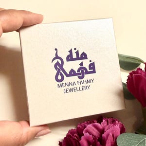 May include: A square, off-white jewelry box featuring a purple logo with Arabic calligraphy and the text Menna Fahmy Jewellery printed in dark purple. The box has a smooth, matte finish and is presented in a clean, simple design.