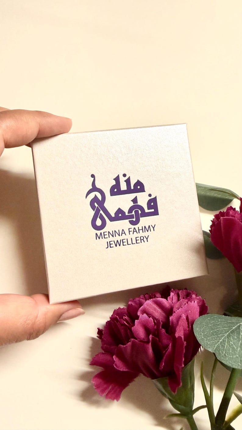 May include: A square, off-white jewelry box featuring a purple logo with Arabic calligraphy and the text Menna Fahmy Jewellery printed in dark purple. The box has a smooth, matte finish and is presented in a clean, simple design.