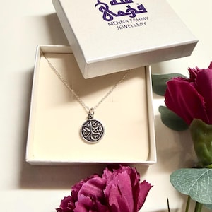 May include: A silver-toned necklace with a circular pendant featuring Arabic calligraphy, presented in a white square box labeled Menna Fahmy Jewellery. The pendant hangs from a delicate chain, and the box lid displays the brand name in purple.