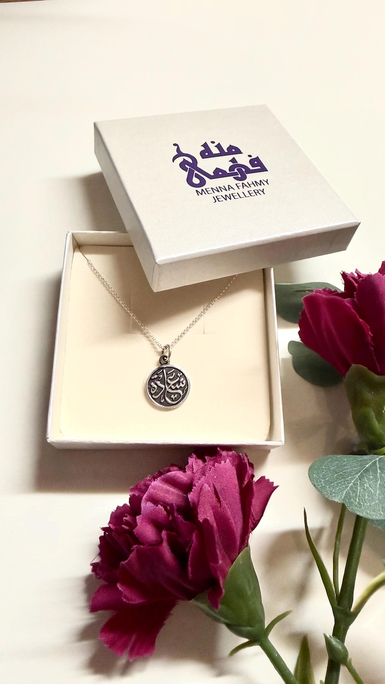May include: A silver-toned necklace with a circular pendant featuring Arabic calligraphy, presented in a white square box labeled Menna Fahmy Jewellery. The pendant hangs from a delicate chain, and the box lid displays the brand name in purple.