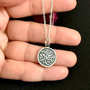 May include: A silver necklace featuring a circular pendant with a dark, recessed background and raised silver Arabic calligraphy. The pendant hangs from a delicate silver chain, showcasing a polished metallic finish and intricate script details.
