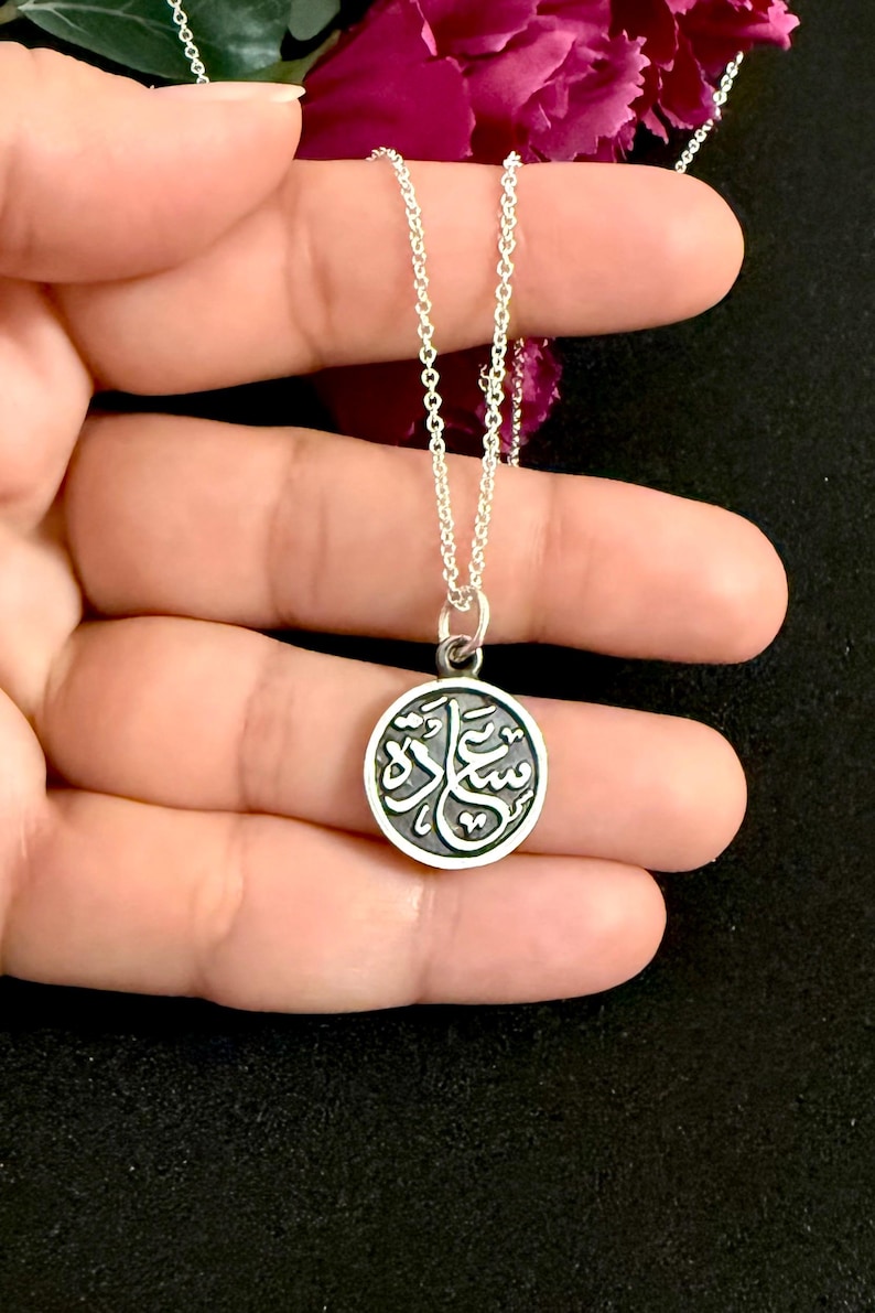 May include: A silver necklace featuring a circular pendant with a dark, recessed background and raised silver Arabic calligraphy. The pendant hangs from a delicate silver chain, showcasing a polished metallic finish and intricate script details.