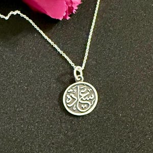 May include: A silver-toned circular pendant necklace featuring Arabic calligraphy on a dark, recessed background. The pendant hangs from a delicate silver-toned chain. The calligraphy is raised and polished, contrasting with the dark metal surface.