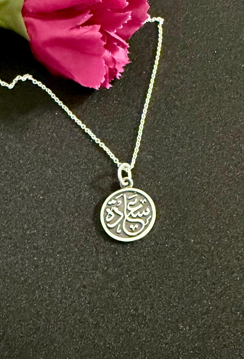 May include: A silver-toned circular pendant necklace featuring Arabic calligraphy on a dark, recessed background. The pendant hangs from a delicate silver-toned chain. The calligraphy is raised and polished, contrasting with the dark metal surface.