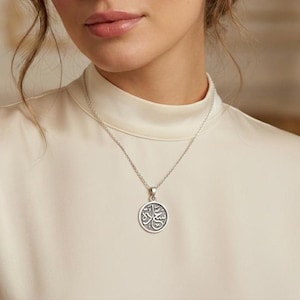 May include: A silver-toned necklace featuring a circular pendant with an engraved Arabic script design. The pendant hangs from a delicate chain, resting against a cream-colored high-neck garment. The metal has a polished finish with dark accents.