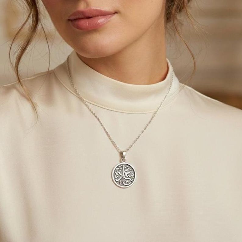 May include: A silver-toned necklace featuring a circular pendant with an engraved Arabic script design. The pendant hangs from a delicate chain, resting against a cream-colored high-neck garment. The metal has a polished finish with dark accents.