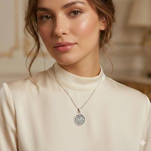 May include: A silver-toned circular pendant necklace featuring an intricate Arabic calligraphy inscription in the center. The pendant hangs from a delicate silver chain, resting against a smooth, cream-colored high-neck satin garment.