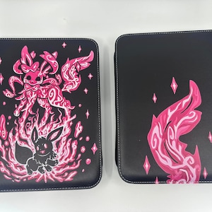 May include: Two black card binders with pink and white designs. One binder features a pink and white creature with a tail and wings. The other binder features a pink and white wing design.