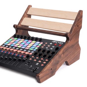 Könnte beinhalten: Ein Ständer aus dunklem Holz mit zwei horizontalen Leisten aus hellem Holz, der einen schwarzen Akai APC40 mkII MIDI-Controller hält. Der Controller verfügt über ein Raster aus bunten quadratischen Tasten, schwarzen Drehreglern und Fadern.