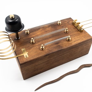 May include: A wooden box with a brass coil, springs, and a bell on top. There are three brass levers on the side of the box. A wooden stick is lying next to the box.