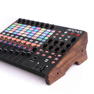 Puede incluir: Un controlador MIDI Akai APC40 mkII con una superficie de control negra, una cuadrícula de botones retroiluminados multicolores, múltiples perillas giratorias y faders, montado en un marco de madera marrón oscuro con veteado.