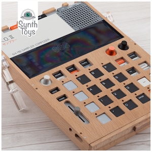 以下が含まれることがあります： 黒と白のディスプレイ、オレンジ色のボタン、グレーのフェーダーが付いた木製サンプラーとシーケンサー。デバイスには「O.II Sampler Composer」と「Synth Toys」のラベルが貼られています。