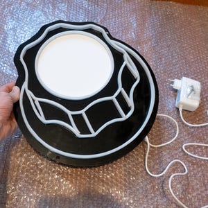 以下が含まれることがあります： 円形で、白いネオンの輪郭を持つ黒と白のネオンサイン。サインは、コード付きの白い電源アダプターに接続されています。