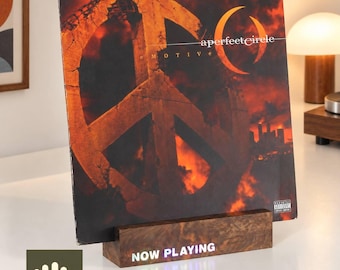 Puede incluir: Una funda de disco de vinilo cuadrada para el álbum eMOTIVe de A Perfect Circle, con un gran símbolo de la paz sobre un fondo naranja y negro ardiente, expuesta en un soporte de madera rectangular con el texto iluminado NOW PLAYING.