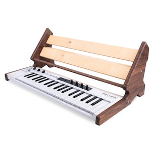 May include: A white Arturia MIDI keyboard controller with black keys sits inside a wooden stand. The stand features dark brown side panels and two light-colored horizontal wooden slats across the back to support the keyboard at an angle.