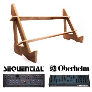 Könnte beinhalten: Ein zweistufiger Synthesizer-Ständer aus hellem Holz. Der Ständer hat ein minimalistisches Design. Darunter befinden sich zwei Synthesizer, einer mit dem Sequential-Logo und einer mit dem Oberheim-Logo.