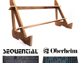 May include: A two-tiered wooden stand for synthesizers, crafted from light brown wood. The stand features a minimalist design. Below, two synthesizers are displayed, one branded with the Sequential logo and the other with the Oberheim logo.