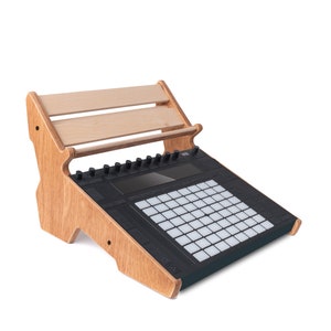 May include: A wooden stand with a shelf for a music production controller. The stand is made of light brown wood and has a black controller with a grid of buttons and a small screen.