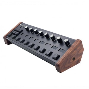 Op de afbeelding: Een zwarte MIDI-controller met een matte afwerking, voorzien van een rij van zeven zwarte draaiknoppen met witte indicatoren en twee rijen vierkante zwarte knoppen. Het apparaat is omlijst door twee donkerbruine houten zijpanelen met zichtbare houtnerf.