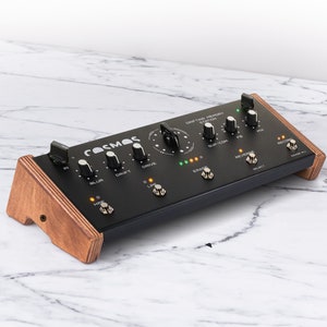 Puede incluir: Un pedal de efectos de audio negro Cosmos Dripping Memory Station con perillas y botones. El pedal está montado en un soporte de madera.