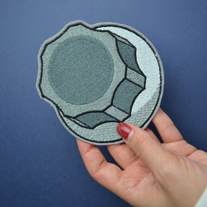 以下が含まれることがあります： 六角形のナットの形をしたグレーの刺繍パッチ。パッチは、隆起したテクスチャデザインとわずかに色あせた外観をしています。