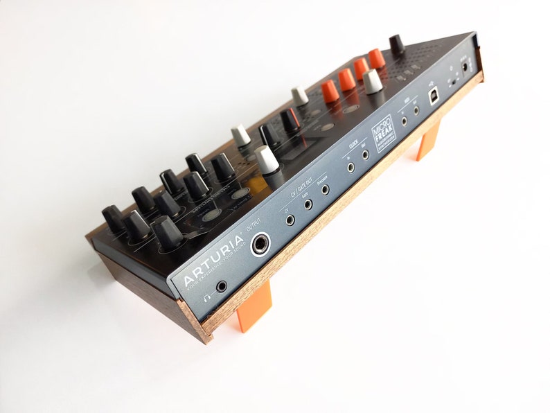 May include: A black and silver Arturia MicroFreak synthesizer with orange knobs and a wooden stand. The back panel shows various ports and connections, including MIDI, USB, and audio.