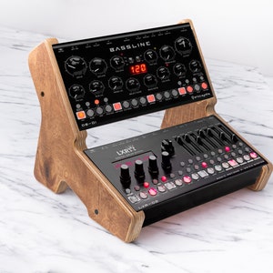 May include: A wooden stand with two black synthesizers. The top synthesizer is a "BASsLINE" with a red digital display. The bottom synthesizer is a "LXRX-02" with a pink digital display.