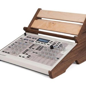 May include: A Waldorf Iridium synthesizer featuring a white control panel with numerous silver knobs, buttons, and a central digital display, mounted on a dark brown wooden stand with two light-colored horizontal wooden support slats.