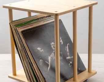 May include: A wooden record storage stand with a light wood finish. The stand has a square top and three vertical supports. The stand is on wheels and has a stack of vinyl records inside.
