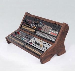 Pode incluir: Um suporte de madeira castanho-escuro contendo quatro sintetizadores Korg Volca. As unidades incluem o Volca Keys, Volca Beats, Volca Sample e Volca Bass, cada um com um painel de controlo preto, vários botões, interruptores e teclado tátil.