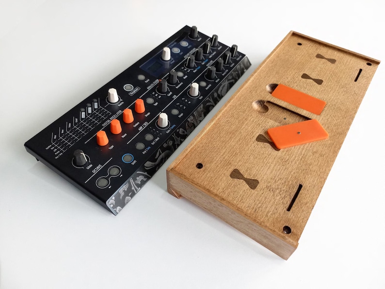 May include: A black synthesizer with orange and white knobs and a wooden stand with orange rubber feet. The synthesizer has a digital display and a variety of buttons and controls.