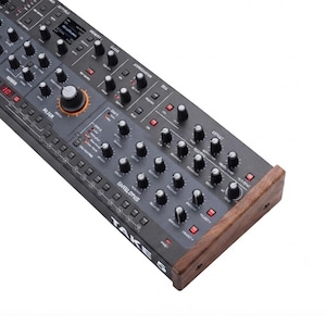 Puede incluir: Un sintetizador gris oscuro etiquetado como TAKE 5 con un panel lateral de madera. El panel de control presenta numerosos mandos negros, pequeños botones rojos y un mando de filtro central grande con un anillo naranja. La interfaz incluye varias etiquetas para la modulación de sonido.