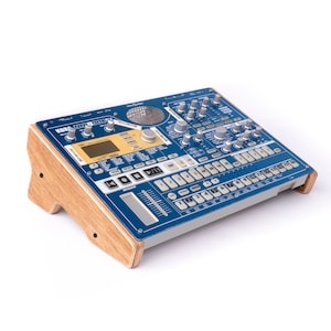 Puede incluir: Un sampler Korg Electribe azul con un soporte de madera. El sampler tiene una variedad de botones, perillas y deslizadores. El texto "KORG ELECTRIBE 2 SAMPLER" está impreso en la parte frontal del sampler.
