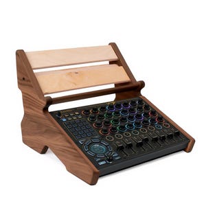 Puede incluir: Un soporte de mesa de mezclas de audio de madera con un panel de control incorporado. El soporte es de madera marrón oscuro con una estantería de madera de color claro. El panel de control tiene numerosas perillas y controles deslizantes con luces de colores.