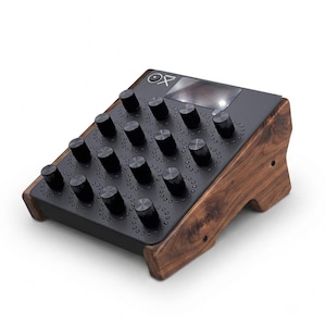 May include: A desktop audio controller featuring a dark matte control panel with fifteen black circular knobs arranged in a grid. The unit is encased in a polished dark wood frame with visible grain and includes a small screen with the logo OX.