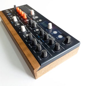 May include: A black synthesizer with a wooden side panel. The synthesizer has a digital display, knobs, and buttons. The text "Cycling Envelope" is visible on the front panel.