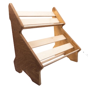 Può includere: Una mensola espositiva in legno a tre livelli con finitura in legno naturale. La mensola ha un design a gradini con tre ripiani orizzontali e uno schienale inclinato. Il telaio è marrone scuro, mentre i ripiani sono di un colore legno più chiaro.