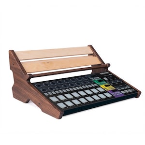 May include: A dark wood and light wood tiered stand holding a black electronic music production controller. The controller features a grid of backlit square pads, multiple circular knobs, and a small digital display screen with various buttons.