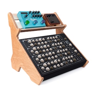 May include: A wooden audio equipment stand holding a black mixer with numerous knobs and buttons. Two effect pedals, one teal and one brown, sit on a shelf above the mixer. The stand is made of light-colored wood.