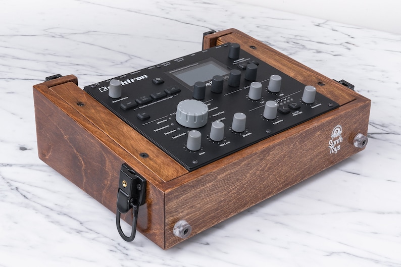 Peut inclure: Un synth&eacute;tiseur de musique &eacute;lectronique noir et gris dans un bo&icirc;tier en bois avec un loquet noir. Le synth&eacute;tiseur a un petit &eacute;cran et plusieurs boutons et molettes. Le bo&icirc;tier porte l'inscription "Synth Toys" sur le c&ocirc;t&eacute;.