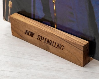 May include: A wooden record stand with the words "NOW SPINNING" engraved on the front. The stand is holding a vinyl record with a person on the cover.