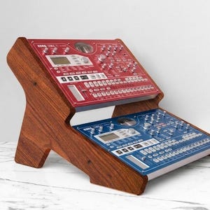 Puede incluir: Un soporte de madera de dos niveles que sostiene una estación de producción musical Korg EMX-1 roja en el estante superior y un sampler Korg ESX-1 azul en el inferior. El soporte tiene un acabado de madera marrón oscuro y soportes laterales.