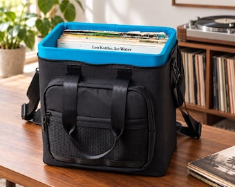 May include: A black square-shaped vinyl record storage bag with a bright blue interior lining. The bag features two top handles, a front zippered pocket with a black mesh overlay, and an adjustable shoulder strap. It is filled with vinyl records.