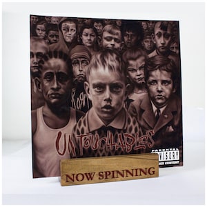 Può includere: Un supporto per dischi in legno con il testo "NOW SPINNING" in lettere rosse. Il supporto tiene un disco in vinile con un'immagine in bianco e nero di un gruppo di bambini. La copertina del disco ha il titolo "Untouchables" in lettere rosse e un adesivo di avvertenza per i genitori.