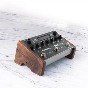 May include: A Strymon TimeLine delay pedal featuring a dark gray metal chassis with black control knobs, a digital display, and three metallic footswitches. The unit is mounted within two dark brown wooden side panels with a natural wood grain.