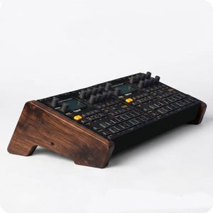 Könnte beinhalten: Ein schwarzer elektronischer Musik-Synthesizer mit zwei digitalen Bildschirmen und zahlreichen Drehreglern und Tasten, inklusive gelber Akzente, montiert auf einem dunkelbraunen Holzständer mit abgewinkelten Seitenteilen für eine erhöhte Position.
