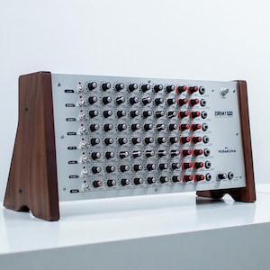 May include: A silver Vermona DRM1 MKIII drum synthesizer with numerous knobs and ports. The device has a rectangular shape with wooden side panels. The controls are black and red. The synthesizer is on a white surface.