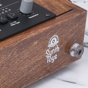 Peut inclure: Une bo&icirc;te en bois avec un panneau noir comportant des boutons et des boutons rotatifs &eacute;tiquet&eacute;s "Fr&eacute;quence", "Type de filtre", "R&eacute;sonance" et "Haut". La bo&icirc;te porte un logo blanc qui dit "Synth Toys" avec une image stylis&eacute;e d'une soucoupe volante.