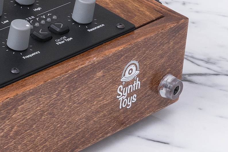 Peut inclure: Une bo&icirc;te en bois avec un panneau noir comportant des boutons et des boutons rotatifs &eacute;tiquet&eacute;s "Fr&eacute;quence", "Type de filtre", "R&eacute;sonance" et "Haut". La bo&icirc;te porte un logo blanc qui dit "Synth Toys" avec une image stylis&eacute;e d'une soucoupe volante.