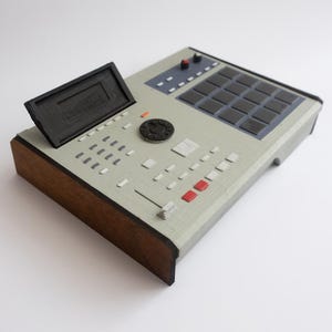 Könnte beinhalten: Ein grau-schwarzes elektronisches Musikproduktionsgerät mit Holzsockel. Zu den Merkmalen gehören ein Display, schwarze Pads, Knöpfe und ein Drehregler. Das Gerät hat eine Retro-Ästhetik.